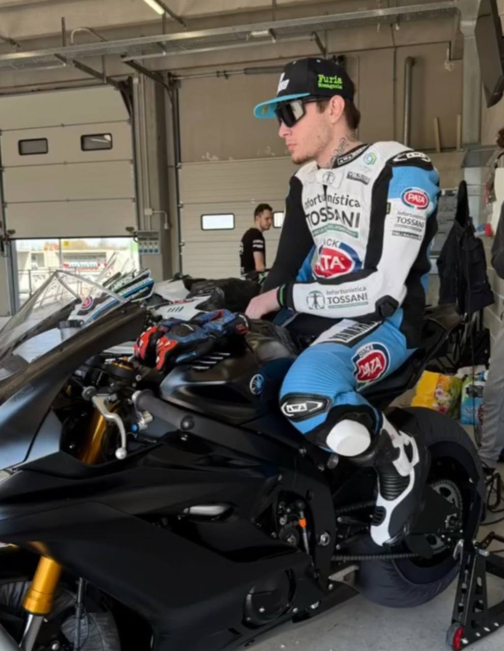Omar seduto su una moto da pista nel box, con tuta, cappellino e occhiali